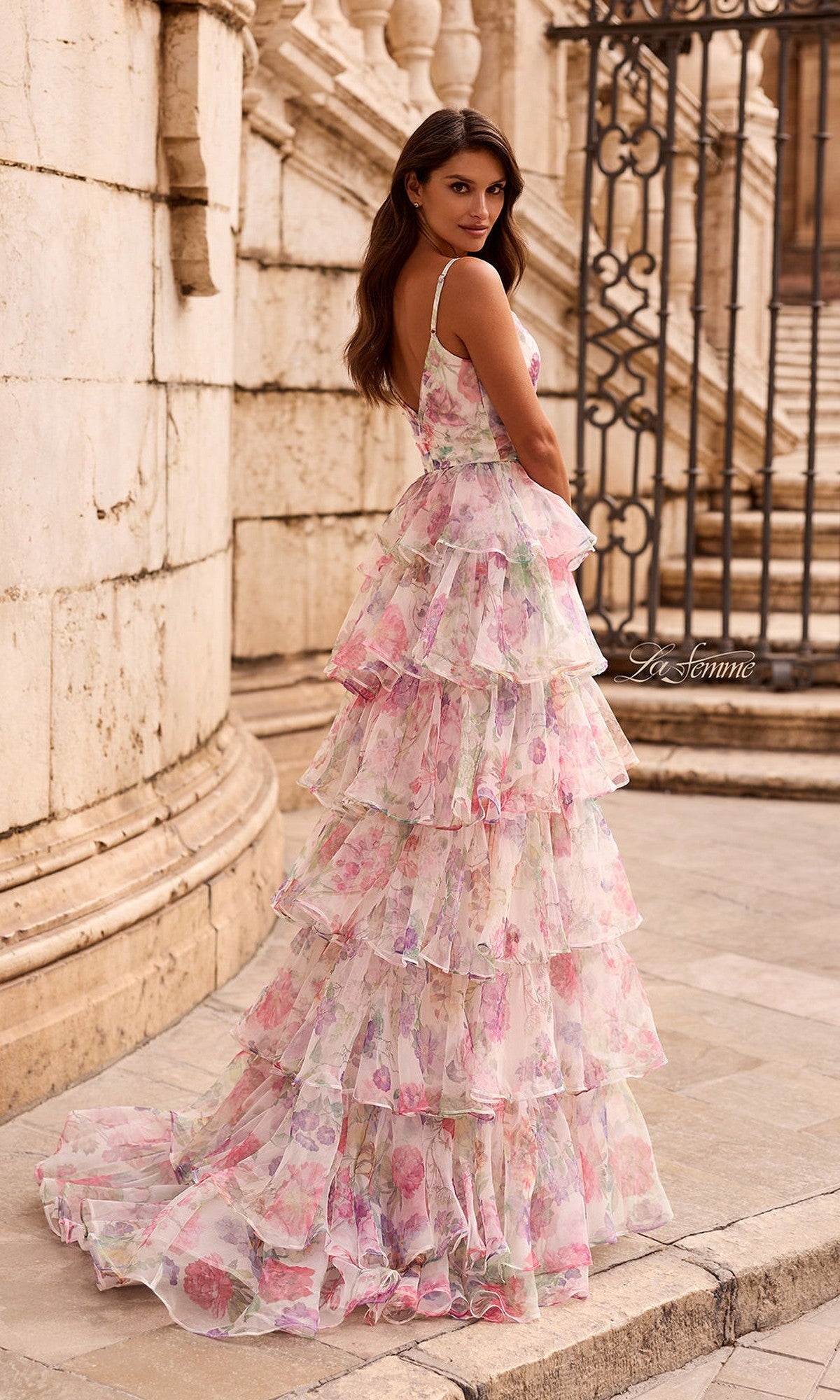 A woman stands outdoors in the La Femme 33740 long prom dress, a chiffon floral gown with tiered ruffles, glancing over her shoulder by stone architecture and a wrought iron gate.